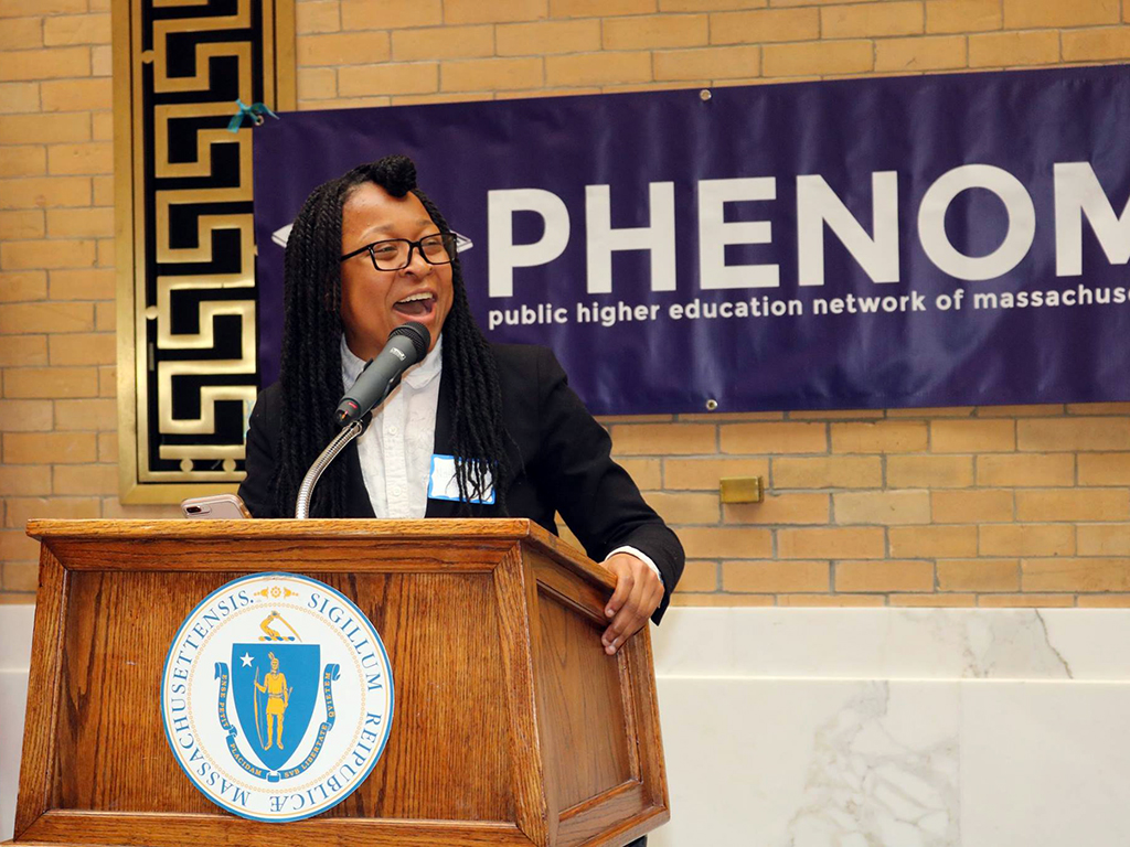 African American female student speaks enthusiastically from podium with PHENOM banner hung behind her